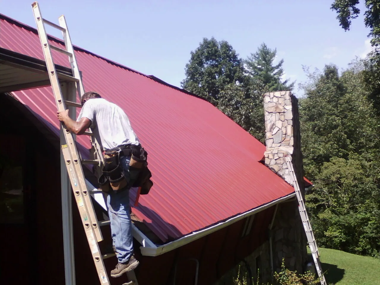 Roofer on a ladder beside a red metal roof and stone chimney — Soffit & Fascia Repair by Roofing Brothers in Max